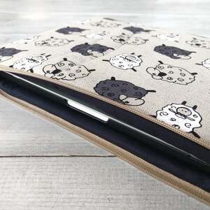 May include: A beige laptop sleeve with a zipper closure. The sleeve is decorated with a repeating pattern of black and white sheep.