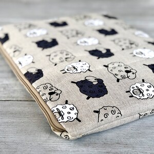 May include: A beige linen zippered pouch with a repeating pattern of black and white sheep. The pouch has a zipper closure and a brown zipper pull.