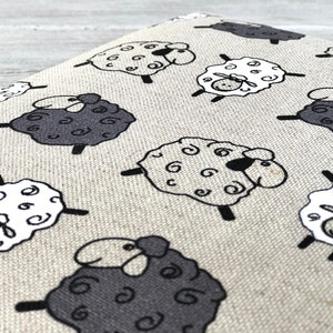 May include: A beige fabric with a repeating pattern of black and white sheep. The sheep are cartoon-like and have curly wool.