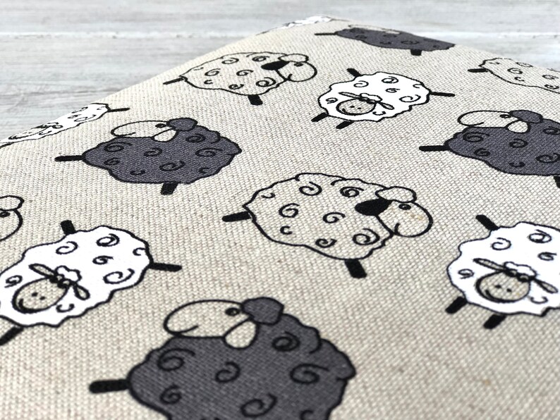 May include: A beige fabric with a repeating pattern of black and white sheep. The sheep are cartoon-like and have curly wool.