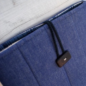 May include: A blue fabric sleeve with vertical stitching, featuring a dark brown rectangular wooden toggle button attached by a black cord loop. The interior lining is visible and displays a blue and white plaid pattern along the top edge.
