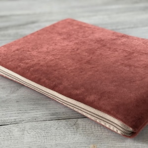 Pode incluir: Uma capa para laptop de veludo rosa empoeirado com fecho de zíper bege. A capa retangular é feita de um material macio e felpudo. O zíper percorre três lados da capa.
