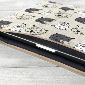 May include: A beige laptop sleeve with a zipper closure. The sleeve is decorated with a repeating pattern of black and white sheep. A laptop is partially visible inside the sleeve.