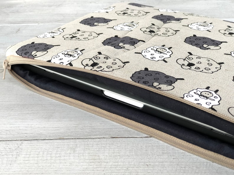 May include: A beige laptop sleeve with a zipper closure. The sleeve is decorated with a repeating pattern of black and white sheep. A laptop is partially visible inside the sleeve.