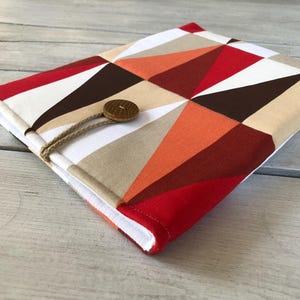 Puede incluir: Una funda de libro de tela con un patrón geométrico y cierre de botón de madera. La funda presenta un diseño de triángulos rojos, blancos, marrones y naranjas. Mide aproximadamente 23 cm por 15 cm.