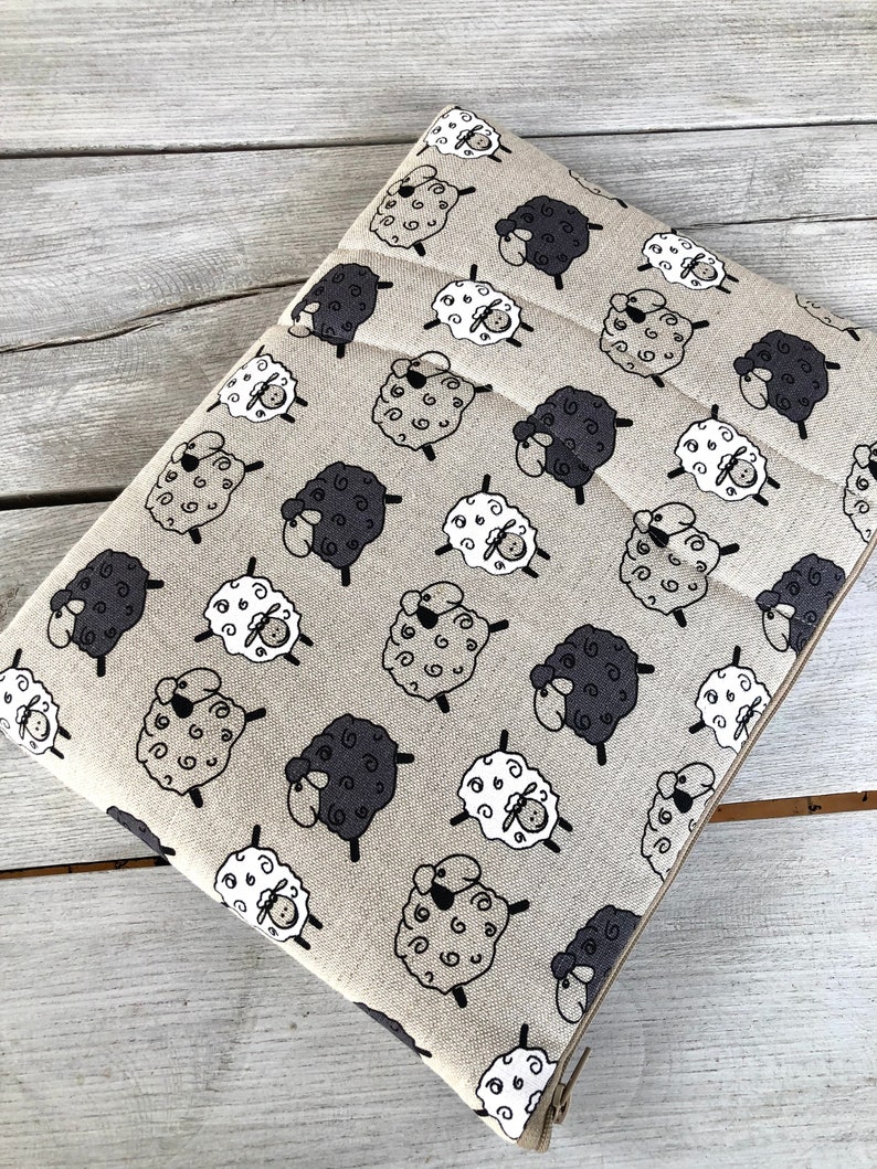 May include: Beige fabric with a repeating pattern of black and white sheep. The sheep are stylized and have curly wool.