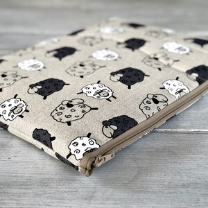 May include: A beige zippered pouch with a black and white sheep pattern. The pouch is made of a soft, textured fabric and has a zipper closure.