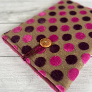Könnte beinhalten: Ein brauner Stoff-Buchumschlag mit einem Muster aus großen rosa und dunkelvioletten Punkten. Der Umschlag wird mit einem Holzknopf und einer passenden Kordelschlaufe befestigt. Der Innenraum und die Kanten sind tief burgunderfarben.