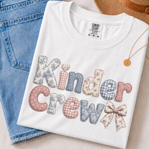May include: A white short-sleeved t-shirt featuring the text "Kinder Crew" in a patchwork-style font with blue and pink floral, gingham, and solid patterns. The design includes a small bow graphic and a Comfort Colors brand label at the neck.