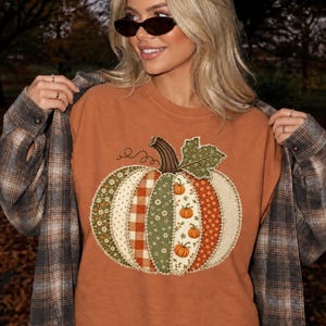 May include: A burnt orange t-shirt featuring a large central graphic of a pumpkin composed of various patchwork-style fabric patterns in shades of green, orange, and cream, layered under a brown and grey plaid button-down long-sleeve shirt.