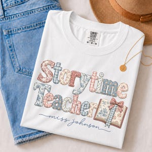 Puede incluir: Una camiseta blanca con el texto Storytime Teacher en un patrón de patchwork floral y de cuadros. El diseño incluye un gráfico de un libro abierto con un lazo. La camiseta tiene cuello redondo y una etiqueta que dice Comfort Colors M-M.