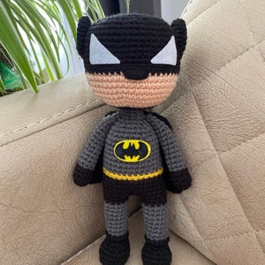 Pode incluir: Um boneco de crochê do Batman com um traje preto e cinza, um cinto amarelo e uma máscara preta com um símbolo branco. O boneco está sentado em um sofá marrom claro.