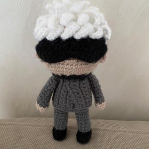 Puede incluir: Una muñeca de crochet con pelo blanco y esponjoso, una máscara para los ojos negra y un suéter y pantalones grises.