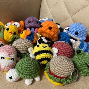 Peut inclure: Une collection de porte-clés animaux crochetés colorés. L'assortiment comprend diverses créatures telles que des hiboux, des pingouins, des abeilles, des tortues et des baleines. Chaque porte-clés est fabriqué avec des coutures détaillées et des couleurs vives.