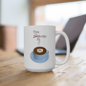 Könnte beinhalten: Eine weiße Keramiktasse mit einem Kaffeetassen-Design. Die Kaffeetasse enthält eine braune Flüssigkeit und ein weißes Schaumdesign mit dem Buchstaben "A" in der Mitte. Der Text "Type JavaScript" ist mit einer roten Linie durchgestrichen und der Text "TypeScript" steht darunter.
