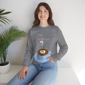 May include: A gray sweatshirt with the text "Type JavaScript?" and a red line through "JavaScript". The sweatshirt also features a blue coffee mug with a white "A" inside, representing the Angular logo.