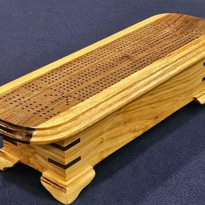 Può includere: Un tabellone di cribbage in legno con finitura marrone chiaro e superficie di gioco marrone scuro. Il tabellone ha una forma rettangolare con estremità arrotondate ed è sostenuto da quattro piedini decorativi. La superficie di gioco è ricoperta di piccoli fori.