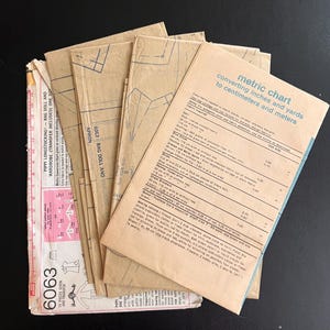 May include: A stack of brown paper sewing patterns for a rag doll and wardrobe. The patterns include a metric chart for converting inches and yards to centimeters and meters. The chart includes measurements for the doll, dress, and other items.