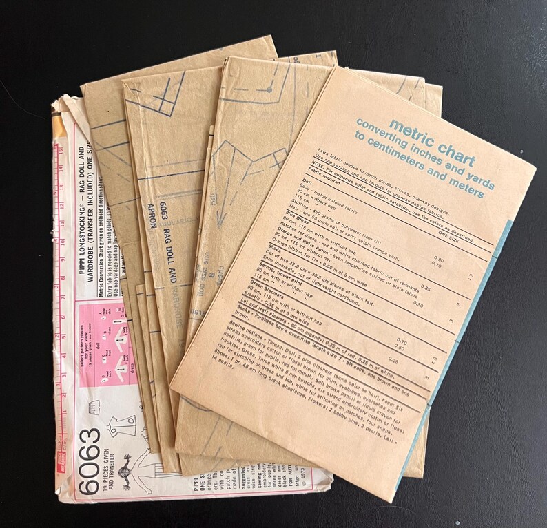 May include: A stack of brown paper sewing patterns for a rag doll and wardrobe. The patterns include a metric chart for converting inches and yards to centimeters and meters. The chart includes measurements for the doll, dress, and other items.