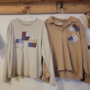 May include: Two long-sleeved shirts hanging on a wooden rack. One is beige with patchwork squares, the other is tan with patchwork squares on the chest. A birdhouse and framed artwork are also visible.