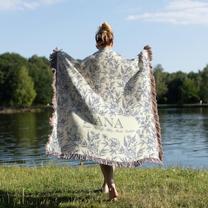 May include: A woman holds a white and blue floral throw blanket with the word "NANA" embroidered on it. The blanket has a fringe border.