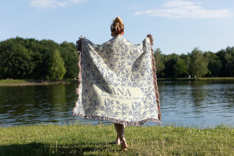 May include: A woman holds a white and blue floral throw blanket with the word "NANA" embroidered on it. The blanket has a fringe border.