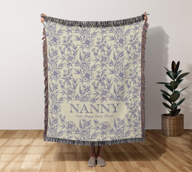 May include: A cream-colored woven throw blanket with a floral pattern in shades of purple. The blanket has a fringe border and the text "NANNY" in a navy blue rectangle with the names "Caleb, Keegan, Linvala, Rowan" underneath.