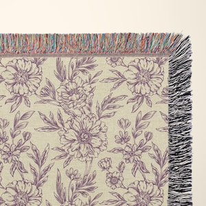 May include: A woven throw blanket with a floral pattern in shades of purple and cream. The blanket has a fringe border.