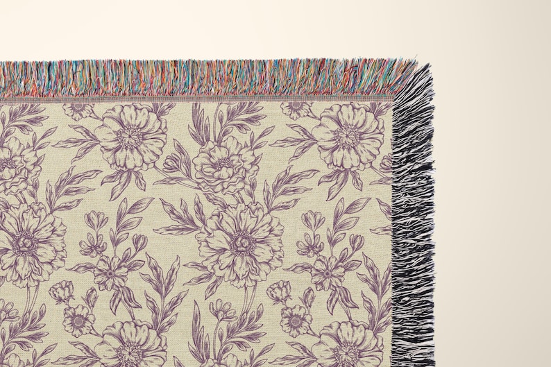 May include: A woven throw blanket with a floral pattern in shades of purple and cream. The blanket has a fringe border.