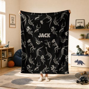 May include: A black rectangular blanket featuring a repeating white pattern of dinosaur skeletons. The name JACK is printed in large white capital letters in the center of the blanket. The fabric has a soft texture and displays various prehistoric fossils.