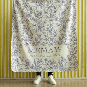 May include: A white throw blanket with a floral pattern in shades of purple. The blanket has the word "MEMAW" printed in black with the names "Michelle, Paul, Lisa, Ashley" printed below in smaller font.