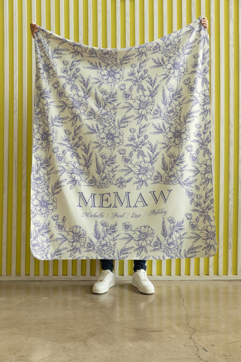 May include: A white throw blanket with a floral pattern in shades of purple. The blanket has the word "MEMAW" printed in black with the names "Michelle, Paul, Lisa, Ashley" printed below in smaller font.