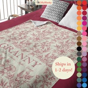 May include: A soft, cream-colored fleece blanket with a pink floral pattern and the word "Granny" in a script font. The blanket is on a red bed with a gray and white patterned pillow.