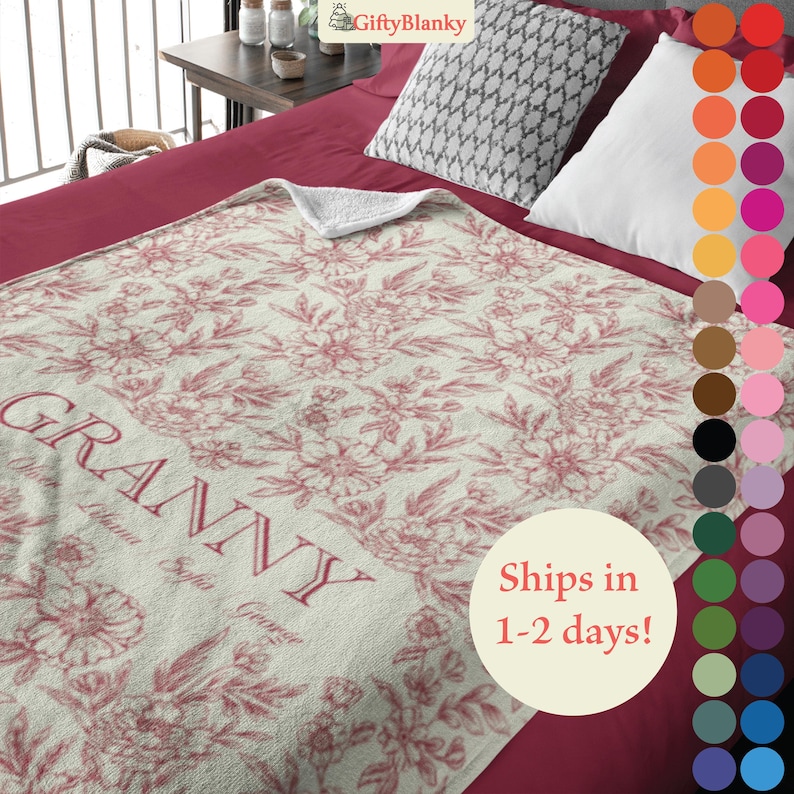 May include: A soft, cream-colored fleece blanket with a pink floral pattern and the word "Granny" in a script font. The blanket is on a red bed with a gray and white patterned pillow.