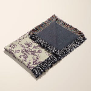 May include: A cream-colored throw blanket with a floral pattern in shades of purple. The blanket has a fringed edge and is folded in half. The back side of the blanket is a solid dark blue color.