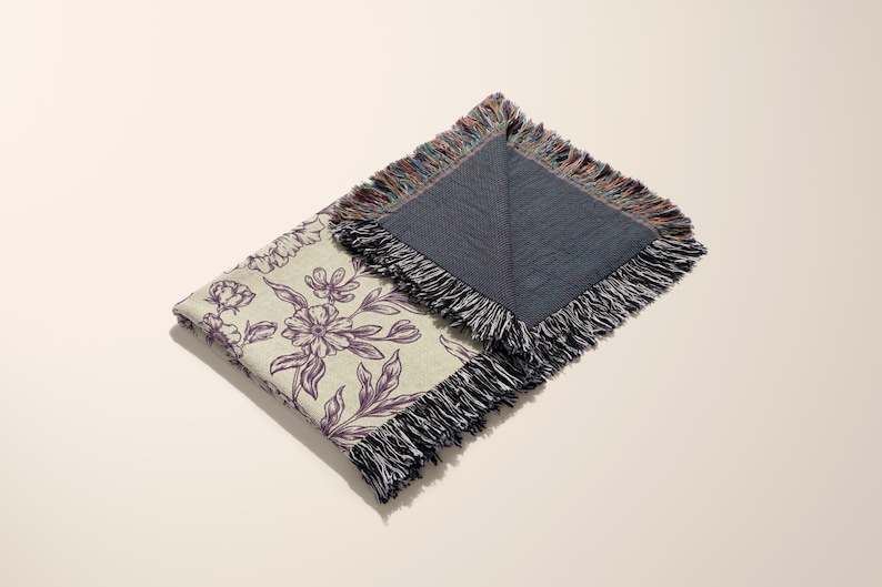 May include: A cream-colored throw blanket with a floral pattern in shades of purple. The blanket has a fringed edge and is folded in half. The back side of the blanket is a solid dark blue color.