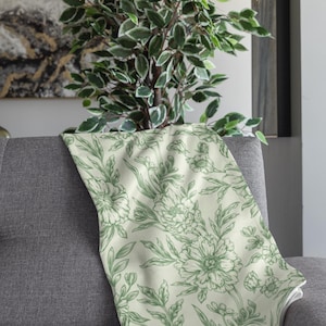 May include: A green floral throw blanket with a white background. The blanket is folded and draped over a gray couch.