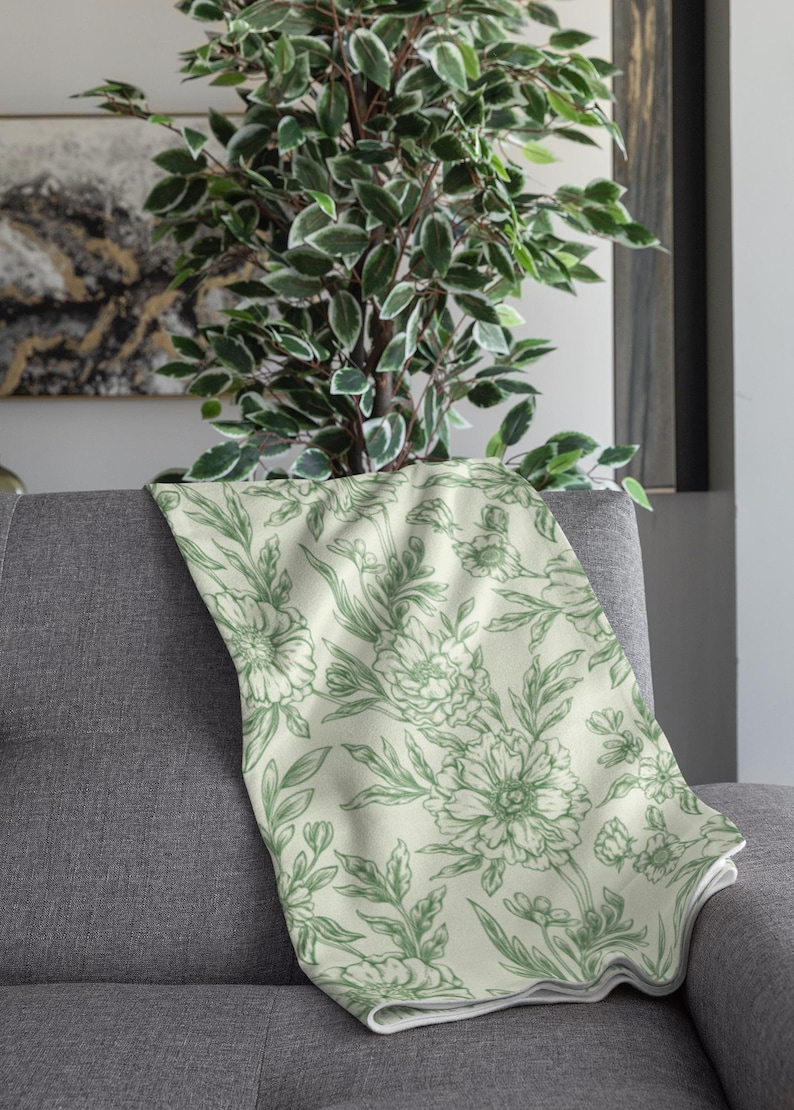 May include: A green floral throw blanket with a white background. The blanket is folded and draped over a gray couch.