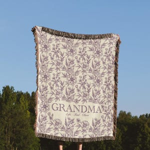 May include: A cream-colored woven throw blanket with a floral pattern in purple. The blanket has fringe on all sides and the text "Grandma" with the names "Lyla, Reed, Oliver" printed in purple.