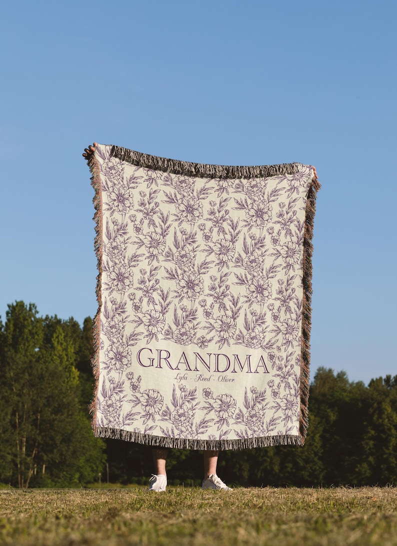 May include: A cream-colored woven throw blanket with a floral pattern in purple. The blanket has fringe on all sides and the text "Grandma" with the names "Lyla, Reed, Oliver" printed in purple.