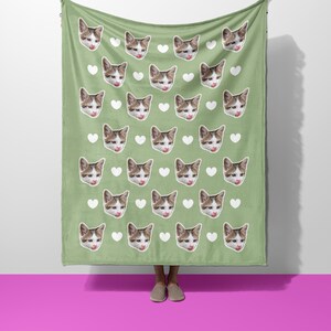 以下が含まれることがあります： 猫の顔と白いハートの繰り返し模様が特徴の緑色のブランケット。猫は茶色と白の毛並みで、舌を出しています。ブランケットは柔らかい素材で作られています。
