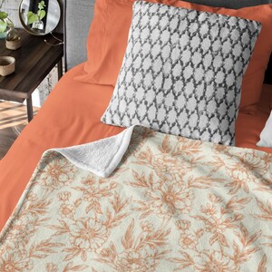 May include: A soft, plush throw blanket with a cream background and a delicate floral pattern in shades of peach. The blanket is folded on a bed with orange and gray pillows.