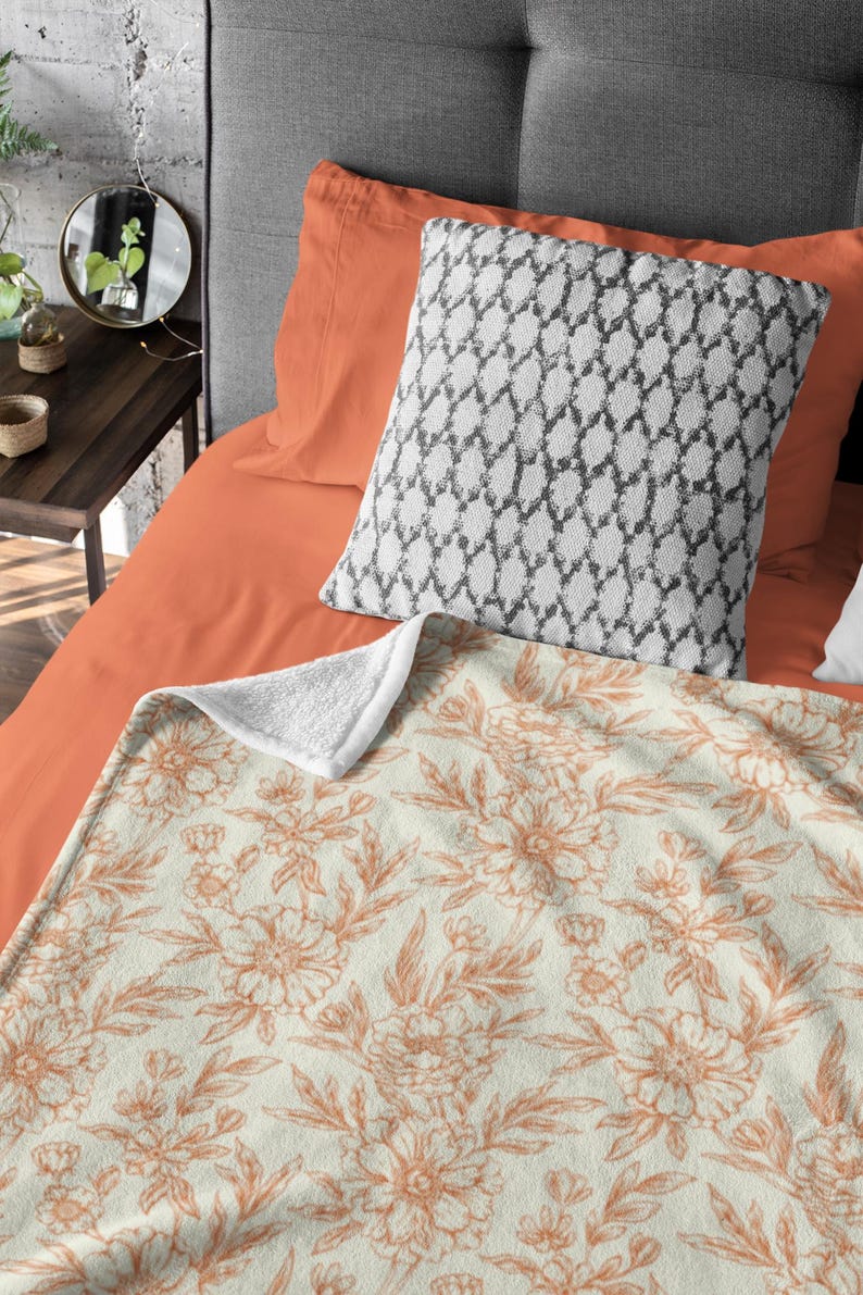 May include: A soft, plush throw blanket with a cream background and a delicate floral pattern in shades of peach. The blanket is folded on a bed with orange and gray pillows.