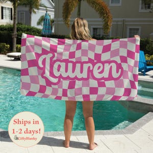 May include: A pink and white checkered beach towel with the name "Lauren" in a cursive font. The towel is held up by a person standing near a pool. The towel has a wavy checkerboard pattern.