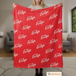 May include: A red blanket with the name "Evelyn" repeated in a white script font. The blanket is held up by a person, showcasing the pattern and color. The blanket is a soft, cozy item.