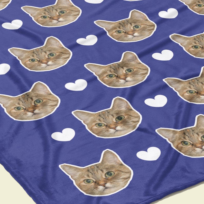 以下が含まれることがあります： 青い毛布。猫の顔と白いハートの繰り返し模様。猫の顔は茶色とタン色で、緑色の目です。毛布は折りたたまれており、模様が表示されています。