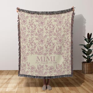 May include: A woven throw blanket with a floral pattern in shades of pink and cream. The blanket has a fringe border and the text "MIMI" in a pink font.
