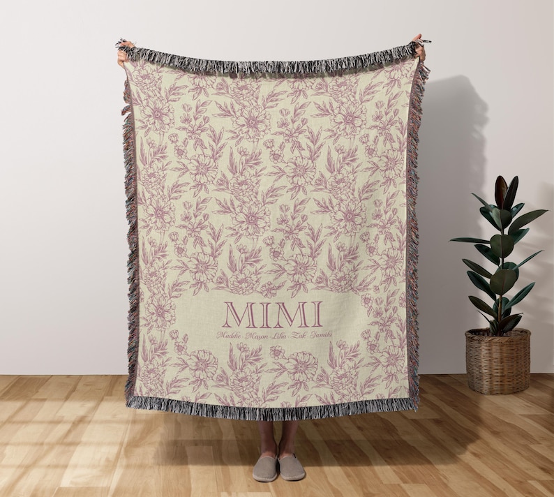 May include: A woven throw blanket with a floral pattern in shades of pink and cream. The blanket has a fringe border and the text "MIMI" in a pink font.