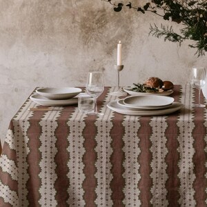 May include: A dining table set for a meal, featuring a patterned tablecloth in shades of brown and cream. The table is set with white plates, glasses, and a vase of flowers. A lit candle adds to the ambiance.