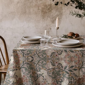 May include: A dining table set with a floral tablecloth in beige, green, and red. White plates, glasses, and a vase of flowers are arranged on the table. A lit candle provides a warm glow to the scene.